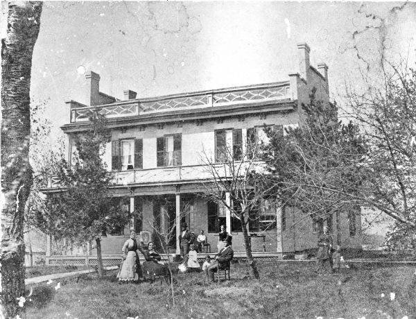 The Abiel E. Brooks House, built in 1853, with a family group posed on the lawn and porch. Identified (front row, left to right) are Mrs. Noah (Jane Ann) Clemmons (standing), daughter, Mrs. Abiel E. (Mary) Brooks and Mr. Abiel E. Brooks, (both sitting). Behind them are Chauncy E. Brooks, Mrs. Manley S. (Julia Maria) Rowley (daughter) and the three Rowley children, Leslie Brooks Rowley, Eugene and Grace. On the far right is Mrs. Chauncy E. Brooks and her daughter, Carrie [?]. Notes written on the negative sleeve indicate that the house was located at 1101 University Avenue at Mills Street and the main entrance was on Mills Street. The block was enclosed by a high board fence.