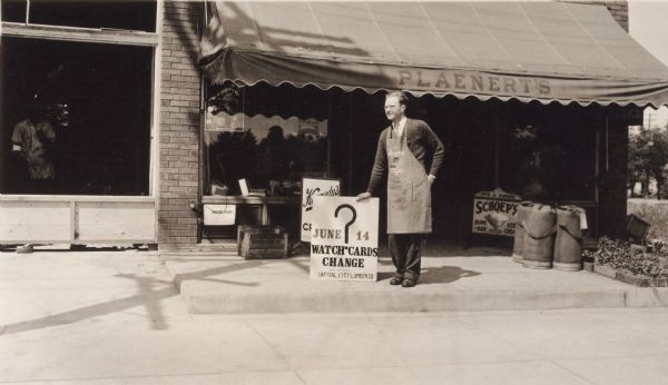 Plaenert's Grocery Walter Plaenert, owner of Plaenert's Grocery, poses in front of his store at 1128 South Park Street. He is holding a sign for the South Side Picnic. The sign reads "June 14, Watch Cards Change, Board Compliments, Capital City Lumber Company." It also has a large question mark in the center. The store has merchandise in the windows and signs for "Kennedy's Creamery," "It's a Food, Schoep's, Home-ade Ice Cream," "Lake Shore Seeds," "Central Wisconsin Co-operative." On the right, vegetables in flats are for sale. On the left is a window with a workman in an apron peering out. His apron is advertising a lumber company. There is a building permit below Plaenert's window.