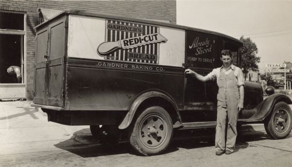 A man dressed in bib overalls poses with a Gardner Baking Company delivery truck. It is parked in front of Plaenert's Grocery. Over the hood in the distance is a Texaco Gasoline sign, underneath it reads "Sanitary Rest Room." Signage on the truck reads, "Gardner's Redi-Cut Bread," "Gardner Baking Co." and "Already Sliced, Ready to Serve."