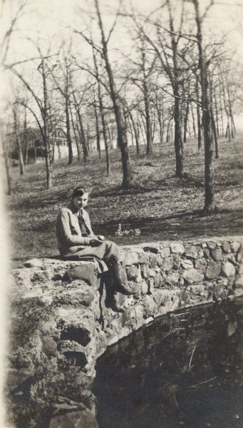 A young woman (possibly one of the O'Dea sisters) sits on the stone wall surrounding the edge of a pond. She is wearing a skirt, blouse, sweater, stockings and shoes, and is holding what appears to be a piece of fruit in her hand. A roll of wire fencing, trees and a house up the hill can be seen in the background.