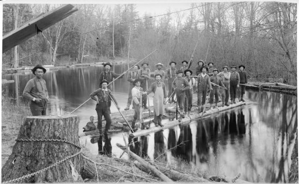 View from shoreline of a driving crew standing on a log raft on the North Branch of the Popple River, a tributary of the Pine River. They are holding long poles, peaveys and cant hooks, tools used to move logs.