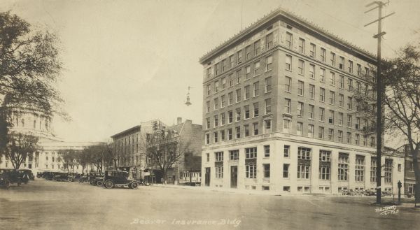The Beavers Insurance Building at the corner of Monona Avenue (currently Martin Luther King, Jr. Boulevard) and Doty Street. It was built in 1921-1922 and designed by local architects, Law, Law and Potter. The Beavers were a fraternal benefit society and, at the time, probably the largest of Madison's home-based insurance companies. The next building to the left is the Orpheum Theatre. On the top of the Orpheum sign it reads, "Vaudeville." The State Capitol appears in the distance on the left. Many automobiles are parked on both sides of Monona Avenue and on the inner curb of the Square. Some construction debris appears along the curb on the right.