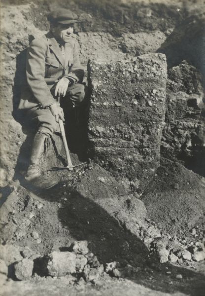Alonzo Pond posing sitting and holding a pickaxe next to a block of earth showing the composition of the escargotiere (an old dump for domestic waste). Here it shows a layer of earth filled with snail shells and artifacts. Pond is wearing a hat, jacket, necktie, jodhpurs and tall lace-up boots.