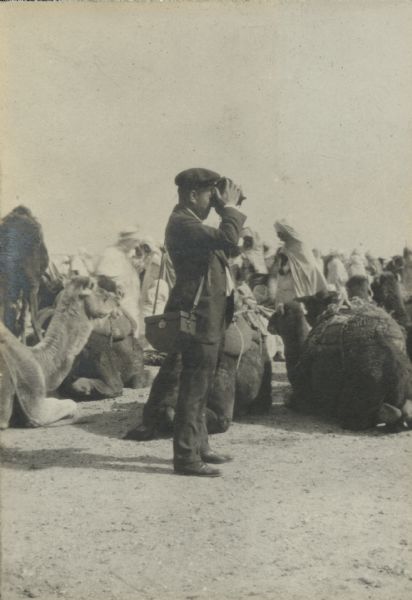 Alonzo Pond using a movie camera at the market. Behind him people are walking among a number of camels sitting on the ground.