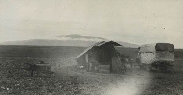 The Pond camp at Medfoun, early in the morning. A tent surrounded by camp furniture and supplies is in the center. A truck is parked beside the tent on the right, and a wheelbarrow is sitting on the left. Clouds and mountains are on the horizon.