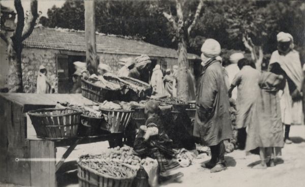 A groups of Algerians shop in a market in Canrobert. Vegetables can be seen in baskets on stepped wooden benches and on the ground. A stone building with a tile roof and trees are in the background.