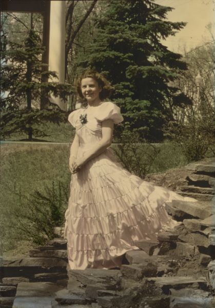 Jean Emily Dyer Reese Outdoor full-length portrait of Jean Emily Dyer (Reese) in her prom dress with a corsage. She is posed on stone steps with the train of her gown spread out behind her and her hands clasped in front. In the background are trees and the columned porch of a building.