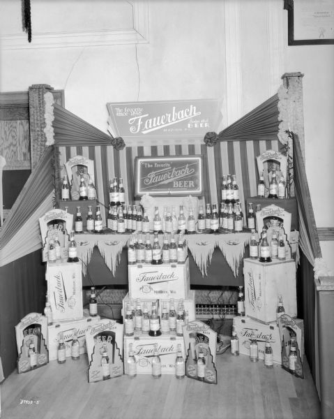 The Fauerbach Beer display at the Grocery Convention. The display consists of many bottles of beer, beer cases, die cut cardboard displays, cloth bunting and two signs. One sign in the center has a neon border.