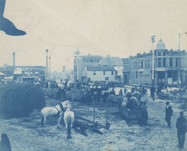 Elevated view of Polish immigrants in Market Square with horse-drawn lumber sledges and hay wagons. Commercial buildings are in the background. A sign on one building reads, "Curran and Wiesner. Kentucky Liquor Store. Bankers, Pawnbrokers."