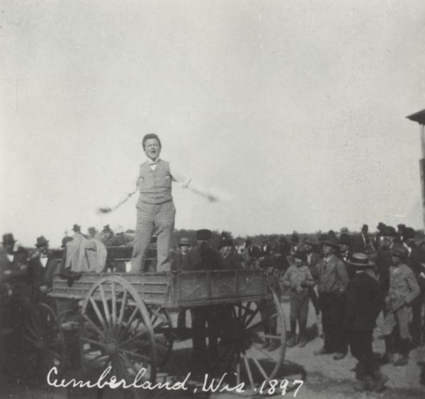 Robert M. La Follette, Sr., with his arms spread wide, speaking from the back of a wagon. It was in part due to his vigorous speaking style that La Follette won the nickname "Fighting Bob." This image is one of a series of views of his appearance at a fair in Cumberland, Wisconsin, in 1897. After three unsuccessful campaigns during which he brought his reform message to Wisconsin at events such as this, La Follette was elected governor of Wisconsin in 1900.