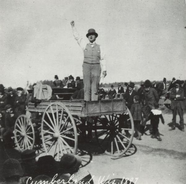 Robert M. La Follette, Sr., with his fist in the air, speaking from the back of a wagon. He is wearing a hat and a suit without the coat. It was in part due to his vigorous speaking style that La Follette won the nickname "Fighting Bob." This image is one of a series of views of his appearance at a fair in Cumberland, Wisconsin, in 1897. After three unsuccessful campaigns during which he brought his reform message to Wisconsin at events such as this, La Follette was elected governor of Wisconsin in 1900.