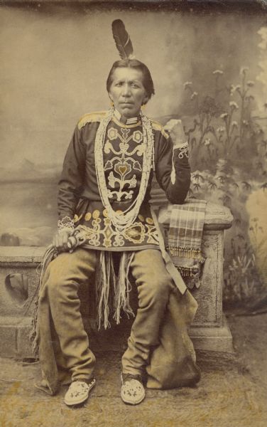 Portrait of a Ho-Chunk Winnesheik Chief Full-length studio portrait of a Ho-Chunk man posing sitting on a prop stone wall in front of a painted backdrop. He is a Winneshiek chief and is wearing Ho-Chunk regalia including embroidered clothing, necklaces, moccasins, and a feather in his hair.