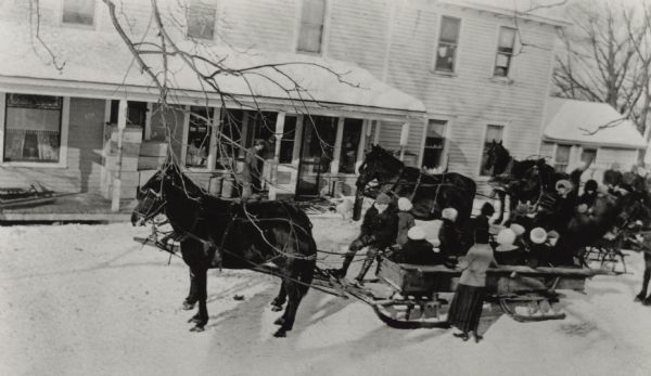Elevated view of a group of people posing on and around horse-drawn sleds. There is a man on the porch of the building in the background.