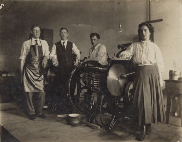 Chandler & Price Platen Press Three men and one women are posing with two Chandler and Price Platen Presses for letterpress printing. The safety guards have been removed from the gear wheels. The woman is wearing a skirt and blouse. One man is wearing a suit with the jacket off with just the vest showing. The other men are wearing work clothes with their sleeves rolled up, and one man on the left is wearing a printer's apron. A spittoon is on the floor in front of the press and on the right is a table with supplies.