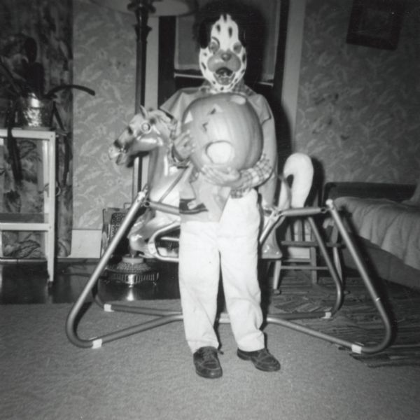 One of Lewis Arms's children (most likely Paul Arms) is wearing a Halloween mask and holding a jack-o-lantern. He is standing in front of a rocking horse on springs.