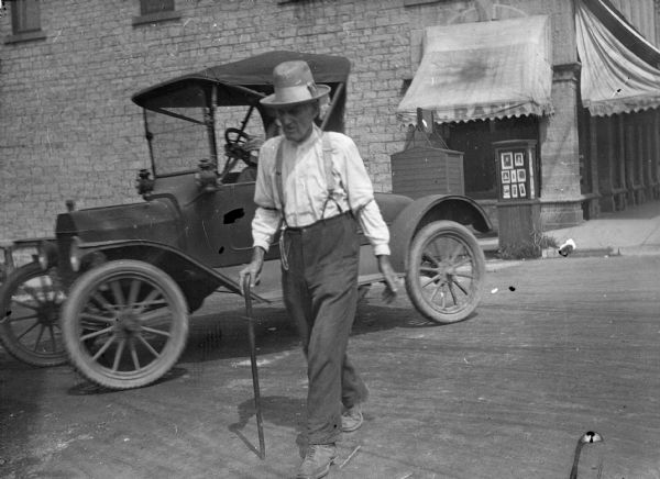 Man Walking with Cane A man walks across the street with the aid of a cane. An Model T style automobile turns the corner behind him. In the background is a business, the awning reads: "A. Rank." An advertising kiosk displays photographic portraits.