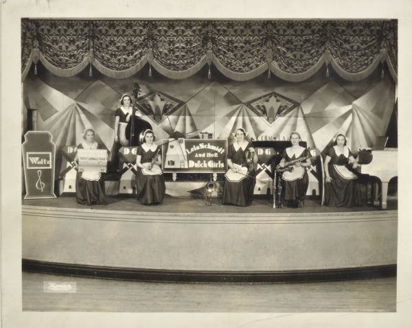 Photograph submitted to the state of Wisconsin for trademark registration. Group portrait of the six women in the Dutch Girls Orchestra. They are posed onstage wearing costumes and holding musical instruments. In the middle of the stage is a sign with a windmill which announces: "Lela Schmidt and her Dutch Girls."