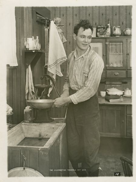 Film still of actor, interior kitchen of the farmhouse. Background includes a Hoosier cabinet.
