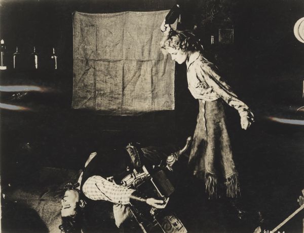 Film Still from "The Boss of the Lazy 'Y'" Actress Josie Sedgwick as Betty Clayton standing over a man laying on his back on debris on the floor. She holds a bottle in her hand, arm raised above her head, poised to hit the man.