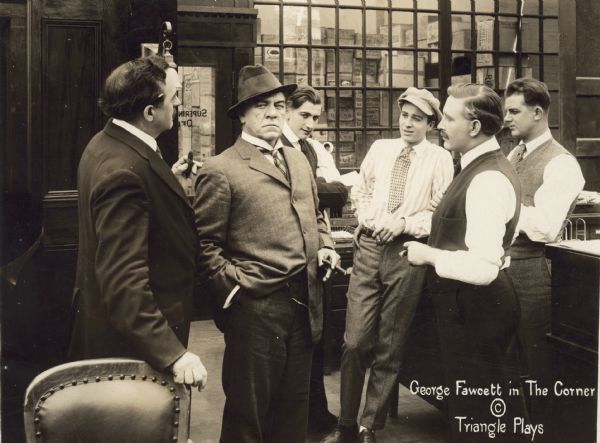 The millionaire David Waltham (played by George Fawcett, holding a cigar and scowling) is in a group of businessmen in an office for a scene still for the 1916 silent drama "The Corner."