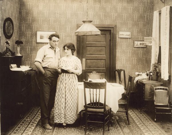 The unemployed worker John Adams (played by Willard Mack) and his wife (Clara Williams) are faced with bills they cannot pay in a scene still for the silent drama "The Corner." They stand in a modest dining room with a birdcage and treadle sewing machine.