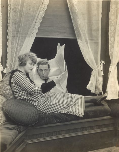 Film still - interior with an actress on a cushioned window seat holding an open newspaper.  An actor is leaning inside the open window, his head coming through the newspaper.