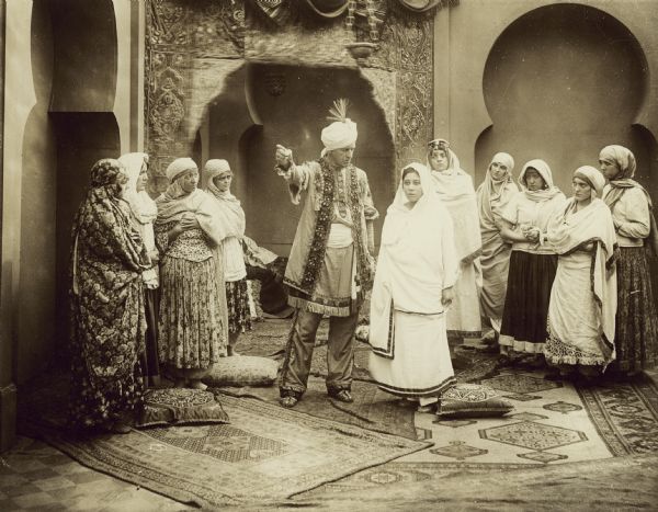 Film still of interior scene, exotic atmosphere, with actor and group of actresses, all in Indian costume with turban.