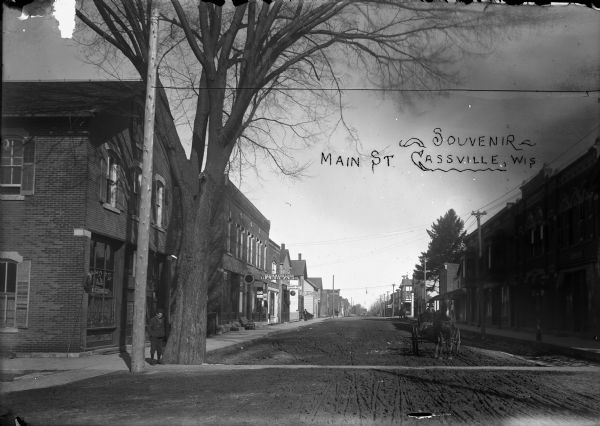 A view of downtown intersection. The view shows the Tate and Kleinpell Hardware store, Dr R.W. Scott, dentist, the Braun Bros with Dubuque Beer on tap. There is a young boy on the sidewalk on the left, and a man in a horse-drawn buggy in the dirt street on the right. Caption read: "Souvenir."