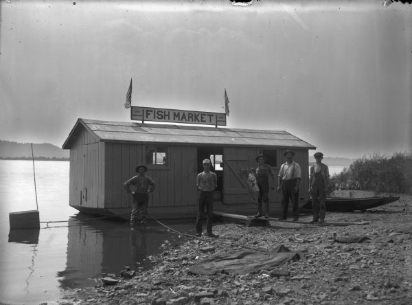 Five men posing in front of the floating fish market, which is on the shore of the Mississippi River. One of the men is holding a large buffalo fish. The man on the left is wearing waders and standing in the water.