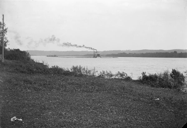 View from shoreline of a steamboat and a long raft or barge far out on the Mississippi River. Smoke is streaming from the smokestack of the sternwheeler.