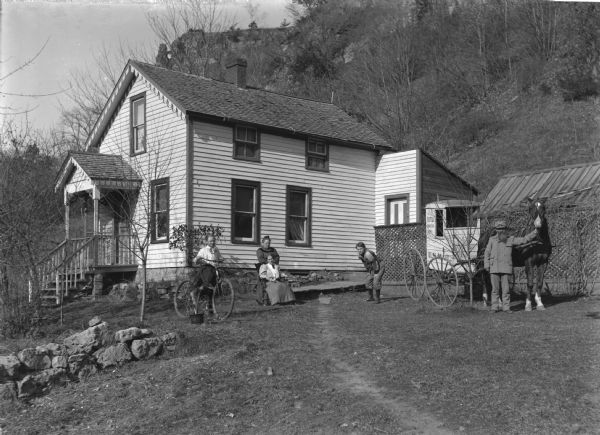 On the right a mailman is standing with his horse and cart. The cart is labeled "RFD Route No.1" and "U.S. Mail." Members of his family, one boy and one girl, and two women, are posing in the yard in front of their house. The young girl is holding a bicycle. The young boy is wearing a football uniform. A tall bluff rises steeply behind the house.