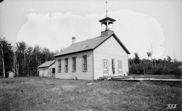 Exterior view of the district school house property, with a young boy standing on the front steps. There is a hand-pump on a wood platform in front of the building.