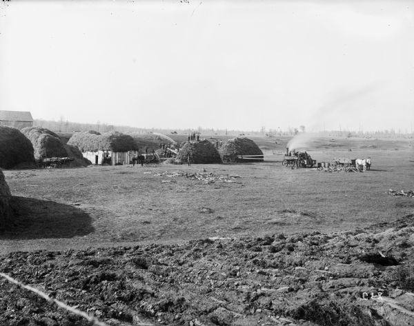 Elevated view from hill of a large group of men harvesting a field. Four men are standing atop a mound of what appears to be straw or hay. A steam thresher, stump puller, barn, and team of horses are in the background.