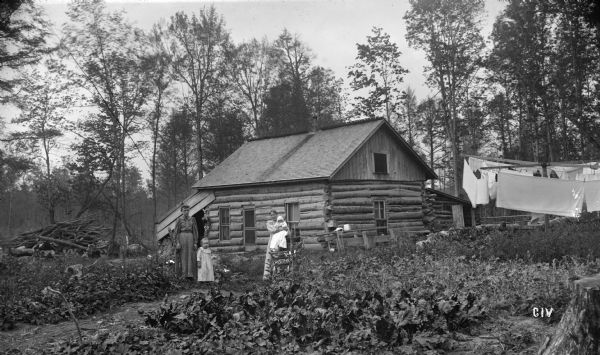 Log cabin of N.G. Willard on 80 acres of land. Two women, a young child, and an infant are posing outdoors in the yard. Laundry is hanging on lines on the right. Trees surround the log house.