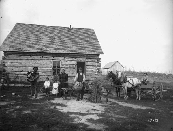 The family of French-Canadian immigrant Peter Lagacy, posing with produce, including a large cabbage, potatoes, and carrots, in front of their log farm home.