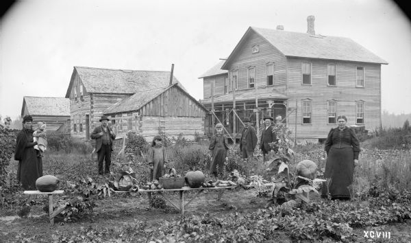 Michael Baltus family posing in front of their log cabin two miles east of Auburndale Station, Wood County, with a new frame house under construction nearby. The table in the foreground is displaying vegetables grown on the farm.