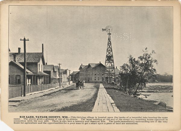 A promotional card displaying a populated village street near a lake in Northern Wisconsin.<p>The caption bellow the photography reads: "RIB LAKE, TAYLOR COUNTY, WIS. — This thriving village is located upon the banks of the beautiful lake bearing the same name. The above gives a glimpse of one of its streets. The large building at the end of the street is a boarding house operated in connection with the saw mill. There is also here a tannery and charcoal kiln. The lands immediately surrounding are of the very finest for agriculture and the opportunities for the poor man to get a start upon a piece of the land are excelled.</p>