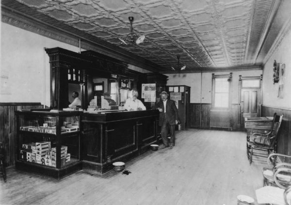 Interior of a saloon. A bartender stands behind the bar, and another man wearing a hat stands in front of the bar. The back of the carved wood bar is mirrored and shows the reflection of the bartender and a large cash register. A calendar hangs on the wall in the background. A glass case on the left side of the bar is filled with tobacco products for sale. The right side of the image shows a desk and chairs lining the wall.