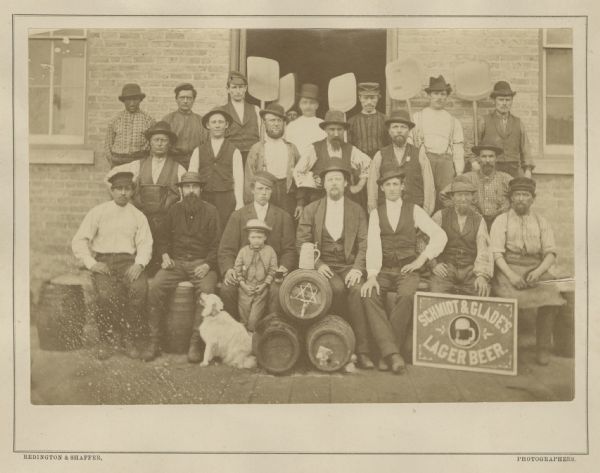 Group portrait of workers, a young boy and a dog outside the Schmidt and Glade Brewery posing with tools and barrels.