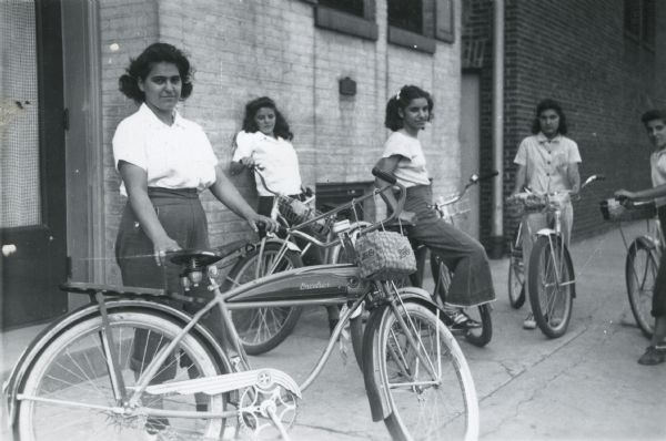 Anita Genna, Florence Quartuccio, Lily Caruso, Francis Musachia [Musachio], and Ann Vitale standing with or sitting on their bicycles preparing to depart from Neighborhood House at 768 W. Washington Avenue on a bike excursion/wiener roast.