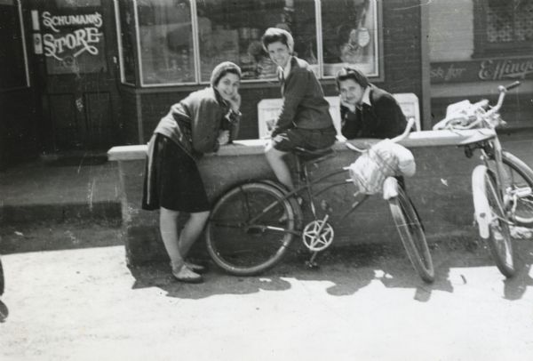 Theresa Manderino [Mandarino?], Vincenza Raimond, and Mary Baldarotta [Baldarotto?] resting and posing on a low wall in front of Schumann's Store, as part of a bike hosteling trip sponsored by Neighborhood House.