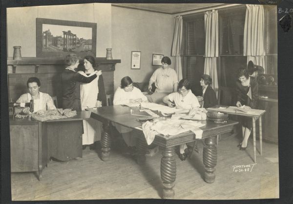 Page from a scrapbook kept by Neighborhood House, with a group of women cutting patterns, sewing by hand and machine, pressing a garment, and checking the fit of a dress in progress.