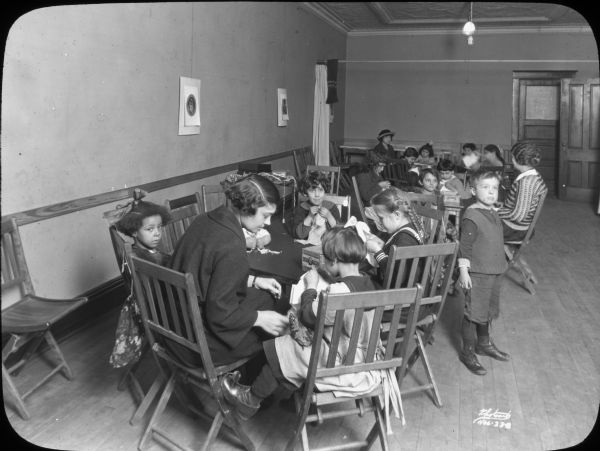 Groups of children are sitting at tables for a Saturday morning sewing group at Neighborhood House, with a woman helping the children at each table. One boy is standing and looking off to the side. A girl sitting in the foreground appears to be looking at the camera.