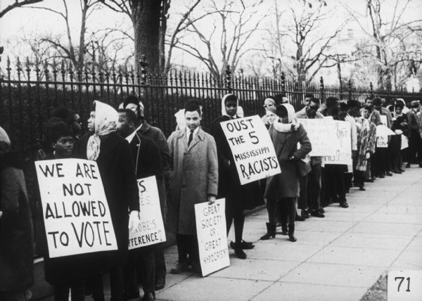 A long line of protesters standing on a sidewalk hold signs calling for voting rights and civil rights. One sign reads, "We Are Not Allowed to Vote". 