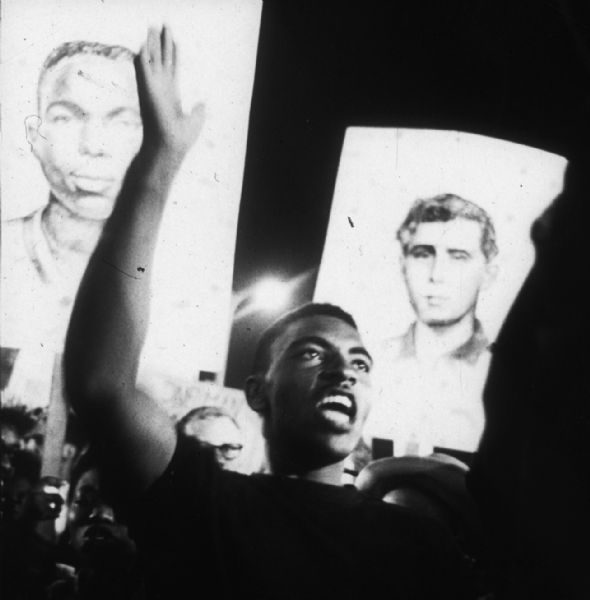 A young man at a nighttime rally (perhaps at a demonstration during the Democratic National Convention in Atlantic City) in support of the MFDP. Behind him people carry placards with portraits of James Earl Chaney and Andrew Goodman, two of the three civil rights volunteers who were murdered during Freedom Summer of 1964.