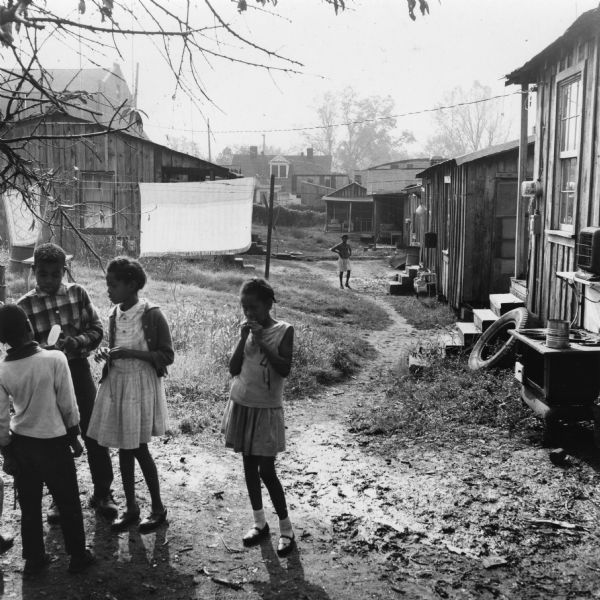 Children gather in the foreground, while houses and yards are visible in the background.  A woman walks along the dirt path towards the camera.<p>“Row upon row of dilapidated buildings, three rows deep, characterize this slum. The city has no housing code and continues to ignore the slums of Natchez. No pavement, no streetlights, 10 outside flush toilets in 2 centrally located shanties shared by 114 people. The 125 persons living in this area share eight water faucets; of these 8, two are inside (installed by tenants) and 6 are outside.”