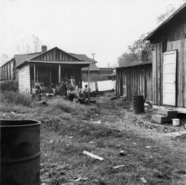 A group of children, several holding balloons, gather on the porch of a building near a debris-scattered yard.<p>“Row upon row of dilapidated buildings, three rows deep, characterize this slum. The city has no housing code and continues to ignore the slums of Natchez. No pavement, no streetlights, 10 outside flush toilets in 2 centrally located shanties shared by 114 people. The 125 persons living in this area share eight water faucets; of these 8, two are inside (installed by tenants) and 6 are outside.”