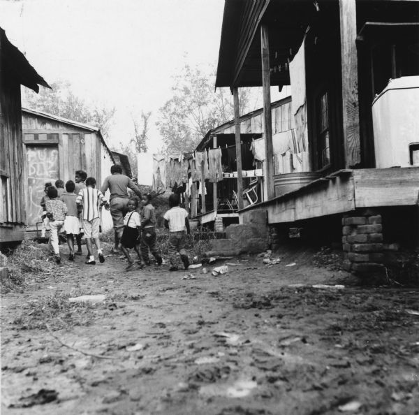 Several children run down a dirt road. Houses are on either side of the road, and laundry hangs from a clothesline above them. The door visible to the left has been painted with graffiti.<p>“Row upon row of dilapidated buildings, three rows deep, characterize this slum. The city has no housing code and continues to ignore the slums of Natchez. No pavement, no streetlights, 10 outside flush toilets in 2 centrally located shanties shared by 114 people. The 125 persons living in this area share eight water faucets; of these 8, two are inside (installed by tenants) and 6 are outside.”