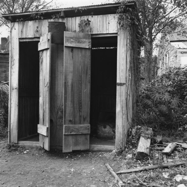 Two of the 13 toilets available in the neighborhood, housed in a small building. Vines grow up the side of the structure and debris is scattered in the surrounding dirt.<p>“Row upon row of dilapidated buildings, three rows deep, characterize this slum. The city has no housing code and continues to ignore the slums of Natchez. No pavement, no streetlights, 10 outside flush toilets in 2 centrally located shanties shared by 114 people. The 125 persons living in this area share eight water faucets; of these 8, two are inside (installed by tenants) and 6 are outside.”