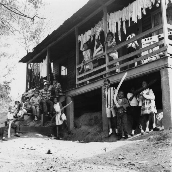 Children stand on the front steps and porch of a home. Many of the children are holding balloons. Laundry hangs from a clothesline on the porch.<p>“Row upon row of dilapidated buildings, three rows deep, characterize this slum. The city has no housing code and continues to ignore the slums of Natchez. No pavement, no streetlights, 10 outside flush toilets in 2 centrally located shanties shared by 114 people. The 125 persons living in this area share eight water faucets; of these 8, two are inside (installed by tenants) and 6 are outside.”